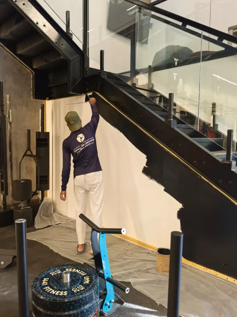 Man painting a wall black underneath a staircase inside a gym.