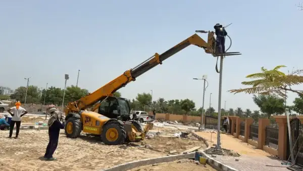 Worker standing in a raised construction lift attached to a JCB machine, spraying or working on a streetlight pole at a construction site.